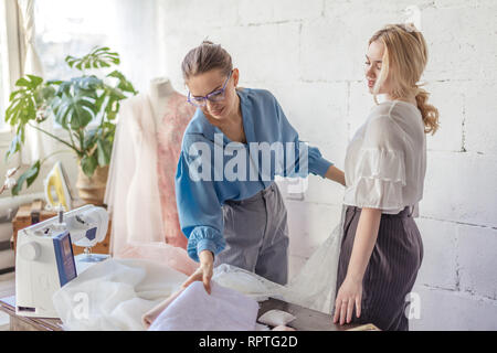 Giovane bella donna bionda sceglie il tessuto per il suo abito da sposa e femmina sarto professionale in Elegante camicetta blu aiuta nel suo Foto Stock