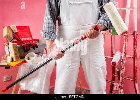 Casa caucasica pittore lavoratore in bianco tute da lavoro, egli tiene nelle sue mani il rullo con estensione. Industria edile. Sicurezza sul lavoro. Foto Stock