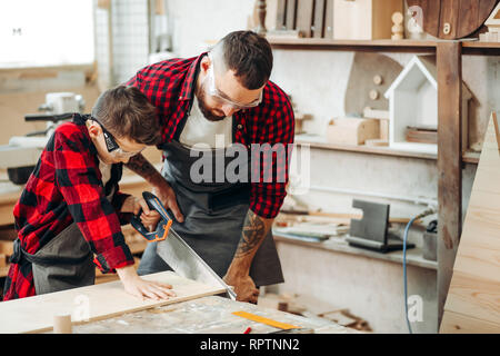 Giovani caucasici padre in occhiali protettivi ad insegnare la sua attenta pensieroso junior school figlio di segare il legno in officina in legno Foto Stock