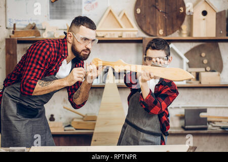 Maschio piccolo falegname creando il suo sogno e il suo papà è aiutare lui.Close-up del giovane maschio carpenter esamina con suo figlio la levigatezza del legno DIY s Foto Stock