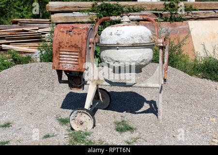 Usato pesantemente sbattuto metallo arrugginito elettrico mescolatore per calcestruzzo al di sopra del mucchio di ghiaia con grandi pannelli in legno e travi in background Foto Stock