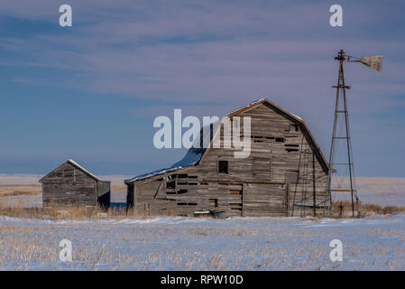 Close up di un fienile vintage, scomparti e mulino a vento circondato da neve sotto una rosa Cielo di tramonto in Saskatchewan Foto Stock