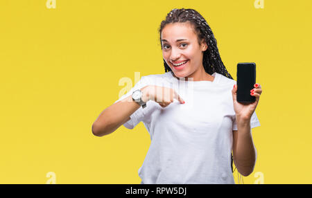 Giovani capelli intrecciati African American Girl mostra schermo dello smartphone su sfondo isolato con sorpresa faccia puntare il dito a se stesso Foto Stock