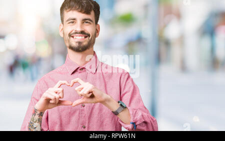 Giovane uomo bello indossare maglia rosa su sfondo isolato sorridente in amore che mostra il simbolo del cuore e la forma con le mani. Concetto romantico. Foto Stock