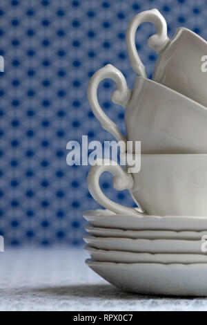 Pila di tazze bianche e piatti su una bianca tovaglia di pizzo. Blu non focalizzato sullo sfondo geometrico. Foto Stock