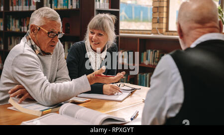 Due alti uomini e una donna seduta in una biblioteca con i libri di corso sul tavolo e discutere. Senior uomini e una donna di condividere idee e discutere di oggetto Foto Stock