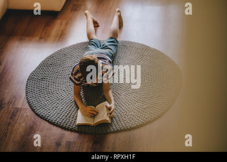 Vista superiore od ragazzo disteso sul pavimento e la lettura del libro. Ragazzo la lettura di un libro a casa. Foto Stock