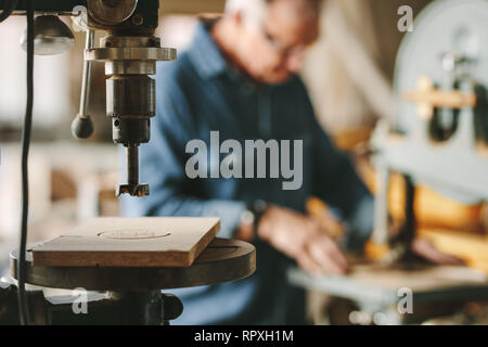 Macchina perforatrice in falegnameria officina con senior falegname lavorando su una sega a nastro in background. Foto Stock