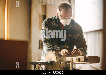 Senior carpenter la levigazione di un legno con levigatrice a nastro in falegnameria officina. Falegname maschio con maschera facciale lavorando sulla levigatrice a nastro macchina. Foto Stock