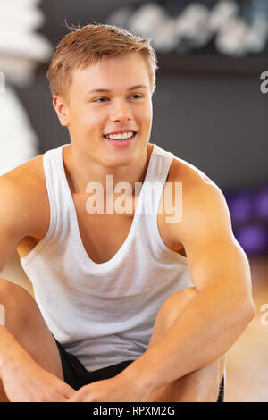 Sorridente giovane uomo vestito di allenamento seduta sul pavimento a palestra per il fitness Foto Stock