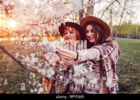 Felice di mezza età madre e figlia abbracciando e tenendo selfie in primavera in fiore giardino al tramonto. La festa della mamma concetto. I valori della famiglia Foto Stock
