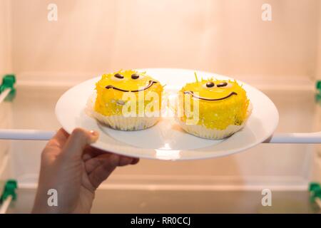Nel frigorifero si mettono due torte fatte in forma di facce con gli occhi e sorrisi. I dolci sono su una piastra bianca detenute da un dolce canto. Foto Stock