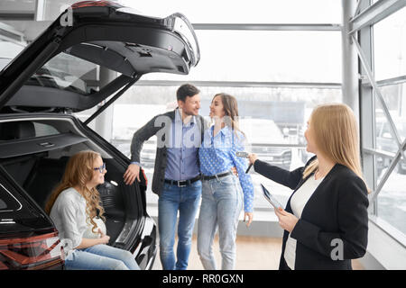 Manager della concessionaria auto di lavoro con i clienti, tenendo la cartella e fornendo chiavi di automobili ad acquirenti di veicolo. Matura in piedi, che pongono, figlia seduta nel baule auto, sorridente. Foto Stock