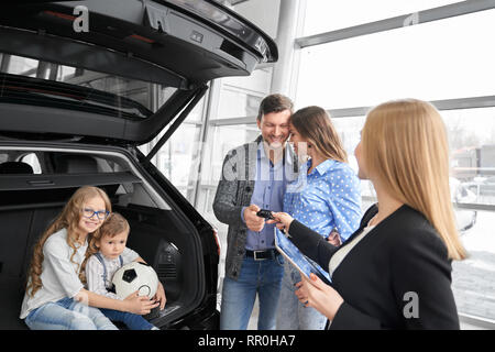 Concessionaria auto della concessionaria showrrom lavorando con clienti, tenendo la cartella e fornendo chiavi di automobili ad acquirenti di veicolo. Matura in piedi, che pongono, la figlia e il Figlio seduto nel baule auto, avvolgente, sorridente. Foto Stock