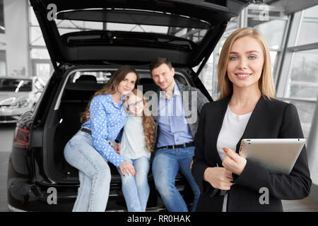 Affascinante donna bionda che lavora in concessionaria auto come concessionario o il manager, donna che guarda la fotocamera, sorridente e tenendo la cartella. La famiglia felice, potenziali acquirenti di auto in seduta baule auto, in posa. Foto Stock