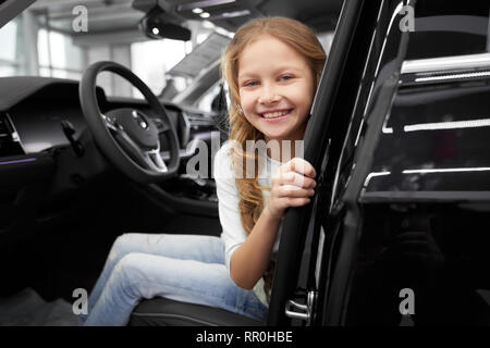 Bello, grazioso piccolo ragazza seduta nel sedile del conducente di nuovo costoso automobile. Incantevole bambino guardando la telecamera, sorridenti e in posa. Concetto di showroom concessionaria. Foto Stock