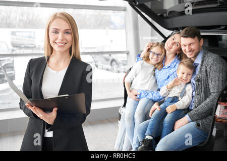 La famiglia felice guardando la telecamera, sorridente e seduta in tronco della nuova automobile. Manager, bella donna che lavorano in concessionaria showroom con i potenziali acquirenti di vetture. Blonde tenendo la cartella in posa. Foto Stock