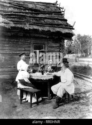 In scena il genere view include l uomo con la balalaika, samovar sul tavolo, ragazzo versando bere, uomo del peering attraverso la finestra e gli altri seduti a tavola al di fuori di un tetto di paglia casa log. Contadina russa s 1875 Foto Stock