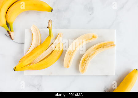 Banane, buccia di banana e una banana pelati tagliati longitudinalmente a metà su marmo bianco bordo e Sfondo marmo. Vista dall'alto. Foto Stock