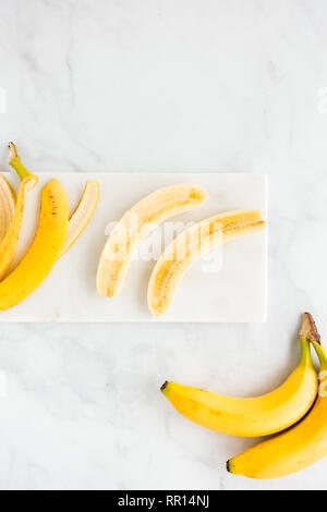 La buccia di banana e una banana pelati tagliati longitudinalmente a metà su marmo bianco bordo e sfondo. Vista dall'alto. Foto Stock