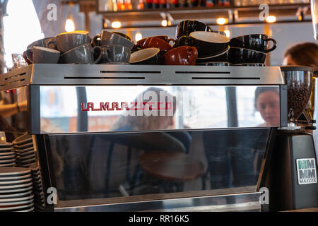 Birmingham, Regno Unito, 24 Febbraio 2019: due donne i capi vengono riflesse in un caffè di argento macchina su un contatore. Le tazze sono impilati sulla parte superiore della macchina. Foto Stock