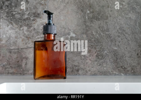 Mano dispenser di sapone in un vetro marrone, bottiglia quadrata, con pompa nero e marmo grigio backsplash vicino al lavandino. Raffiguranti le mani pulite, lavaggio. Foto Stock