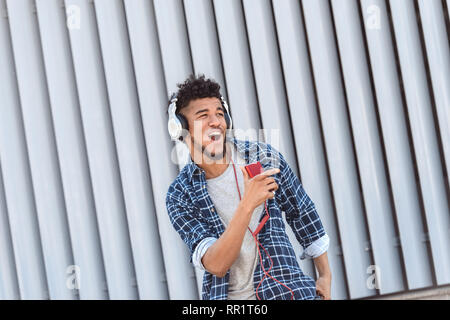 Per il tempo libero all'aria aperta. Mulatto guy nelle cuffie in piedi isolato su una strada di città ascoltando musica canto gioioso Foto Stock