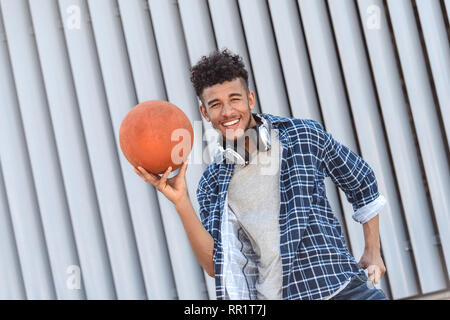 Per il tempo libero all'aria aperta. Mulatto ragazzo con le cuffie sul collo in piedi isolato su una strada di città tenendo palla sorridendo giocoso Foto Stock