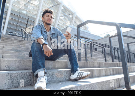 Amanti della musica. Mulatto guy auricolari in seduta sulle scale con lo smartphone ascoltando musica pensieroso vista dal basso Foto Stock