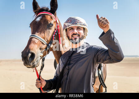 Medio Oriente uomo bello con il tipico abito emirates in sella ad un cavallo arabo nel deserto di Dubai Foto Stock