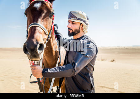 Medio Oriente uomo bello con il tipico abito emirates in sella ad un cavallo arabo nel deserto di Dubai Foto Stock