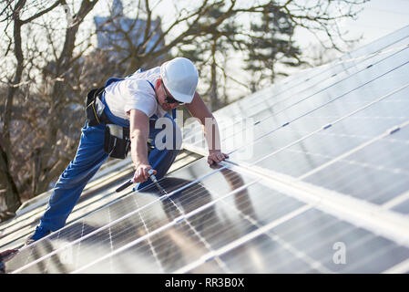 Lavoratore di sesso maschile installazione solare pannello fotovoltaico sistema utilizzando un cacciavite. Elettricista blu montaggio modulo solare sul tetto di casa moderna. L'energia alternativa concetto ecologico. Foto Stock