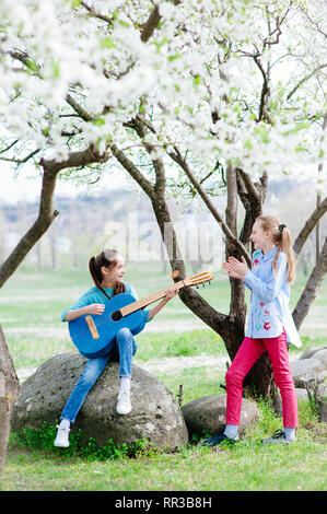 Felice bambine all'aperto le prove con la chitarra e cantare in fiore parco di primavera Foto Stock