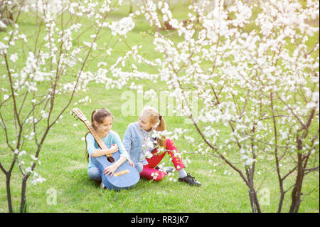 Due sorridenti bambine con la chitarra acustica seduto sul prato verde sotto alberi in fiore in primavera Foto Stock