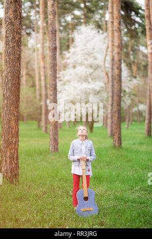 Bellissima bambina con chitarra vintage getting ispirato piedi tra gli alti alberi nel Parco di primavera Foto Stock