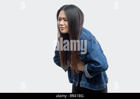 Mal di stomaco. Vista laterale ritratto della bella bruna asian giovane donna casual in denim blu con camicia in piedi e la sensazione di dolore sul suo stomaco. interna Foto Stock