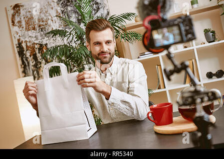 Shopping online. Bello il blogger maschio tenendo la carta shopping bag e guardando la telecamera mentre lo streaming live. Youtube. Blog di moda. Foto Stock