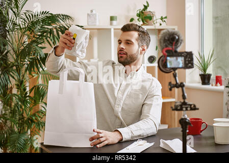 Blog di cibo. Giovani blogger barbuto tenendo fuori un panino durante la registrazione di un nuovo video per il suo canale di Youtube. Concetto di cibo. Gastronomia Foto Stock