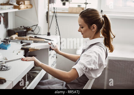 Preparazione di un ambiente di lavoro...vista laterale del giovane femmina gioielliere la preparazione di luogo di lavoro per la realizzazione di nuovi gioielli. Gioielli. Business. Workshop di gioielli. Lavorare pro Foto Stock