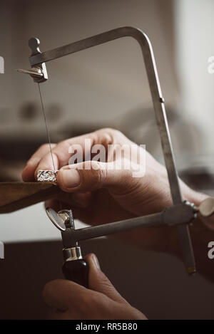 Momento critico. Close-up foto del gioielliere le mani facendo un anello color argento con professional gioielliere regolabile la sega a telaio il suo banco di lavoro Foto Stock