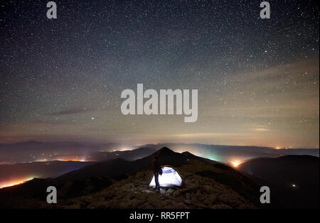 Tourist Camp estivo di notte sulla cima della montagna rocciosa. Silhouette di maschio escursionista appoggiato accanto incandescente tenda sotto la splendida notte cielo pieno di stelle. Sullo sfondo il cielo stellato e città luminosa Foto Stock