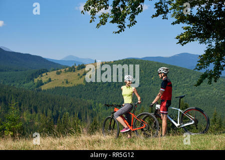 Felice bikers matura, l uomo e la donna in piedi con la bici sulla collina erbosa sotto il grande albero verde ramo, sorridente e cercando di ogni altro su soleggiate giornate estive. Bella vista delle montagne sullo sfondo Foto Stock