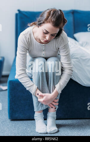 Sconvolto donna abbracciando le ginocchia e seduta sul letto di casa Foto Stock