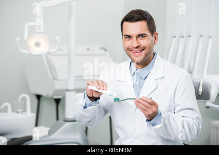 Sorridente dentista spremitura di dentifricio su uno spazzolino da denti in clinica dentale Foto Stock