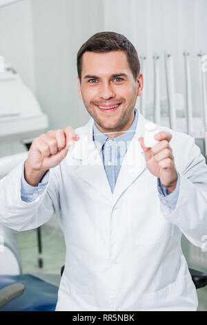 Allegro dentista in camice bianco a sorridere mentre si tiene il filo interdentale Foto Stock