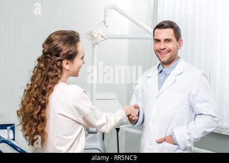 Sorridente dentista in piedi con la mano in tasca e stringe la mano al paziente di sesso femminile Foto Stock