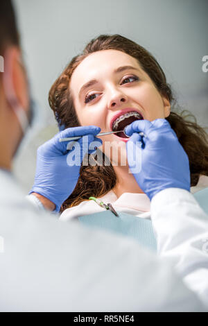 Messa a fuoco selettiva di donna attraente in parentesi graffe durante l' esame dei denti nei pressi di dentista Foto Stock