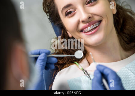 Close up di allegra donna in parentesi graffe durante l' esame dei denti nei pressi di dentista Foto Stock