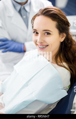 Messa a fuoco selettiva della donna in parentesi graffe sorridente con il dentista in piedi con le braccia incrociate su sfondo Foto Stock