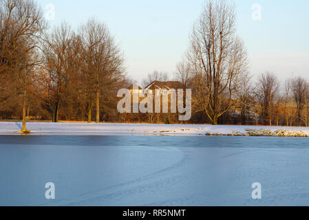 Coperta di neve park il marciapiede, lago di dock e case con fondo in lontananza al tramonto Foto Stock